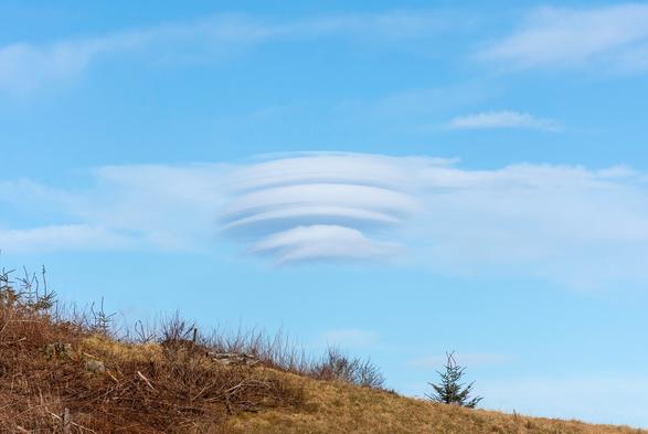 A stack of lenticular, disc-like clouds in a pale blue sky, the near horizon of grass slopes from left to right at the bottom of the shot