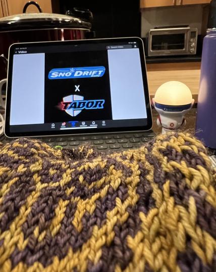 A purple and gold hand knit sweater lies piled in a heap in front of an iPad with a video “Snow drift x Tabor Racing Team” which is next to a small light in the form of an astronaut with a big head; a purple hydro flask coffee mug; and a metallic red crock pot on a butcher block board in the background.