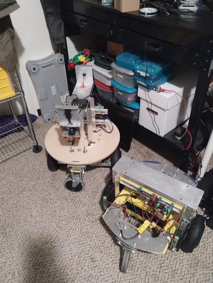 Picture of two custom build hub wheel robots in the fore ground.  
In the back ground a bunch of parts bins sitting on a lower shelve of my work bench.