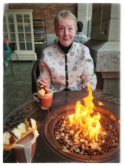 This image was made on the front patio of the Fairmont Empress Hotel. Although it can't be seen in this image, the patio overlooks the Inner Harbour across the street. In this image is a large, round, black metal table with a fire pit in the middle, fed by a gas flame. Across from me, Marcia is looking as beautiful as always, with her gorgeous blue eyes, pixie-cut hairdo and Chinese-style white jacket with a black geometric pattern. Also in the image are two cups of the Fairmont's hot chocolate, with whipped cream around the edge of the glass mugs, and topped with a dog-bone-shaped cookie dotted with icing, red velvet cake and slivers of dark chocolate.