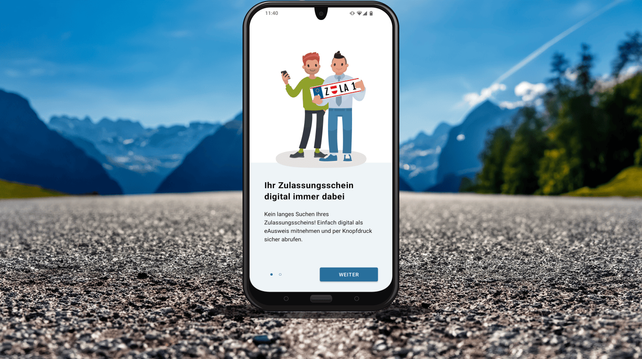 Ein Handy steht auf einer Alpinstraße, darauf zu sehen ist eine Infografik mit zwei gezeichneten Menschen, die ein Kennzeichen halten, darunter steht: Ihr Zulassungsschein digital immer dabei! Kein langes Suchen ihres Zulassungsscheins. Einfach digital als eAusweis mitnehmen und per Knopfdruck sicher abrufen.