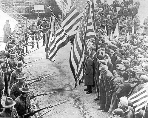 Massachusetts militiamen with fixed bayonets surround a group of strikers. Men and women in the front are holding US flags on long staffs. Everyone is dressed in long, woolen coats against the cold winter. The militiamen are standing with their rifles at their waists, aiming at the protesters. By http://womhist.binghamton.edu/teacher/DBQlaw2.htm, Public Domain, https://commons.wikimedia.org/w/index.php?curid=131378