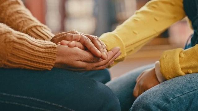 A photograph of two people cropped to show only their thighs and hands. The person on the left is dressed in a brown jumper and dark jeans is holding the hand of the person on the right between both their hands. The person on the right is dressed in a yellow jumper and light blue jeans