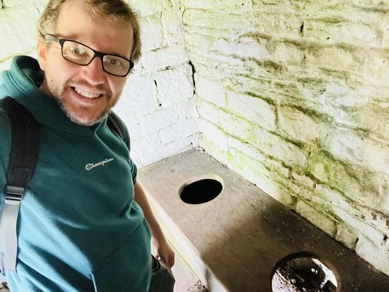 A selfie of the writer in a green hoodie stood next to a double wooden latrine