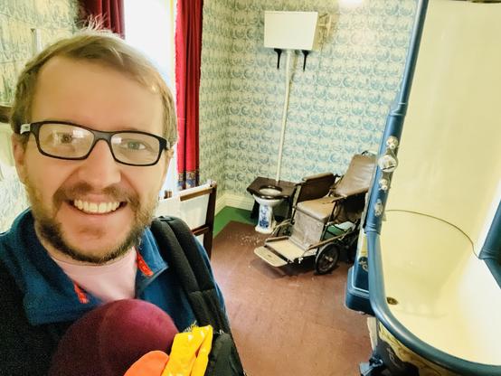A selfie of the writer carrying a baby in a sling in front of a Victorian bathroom