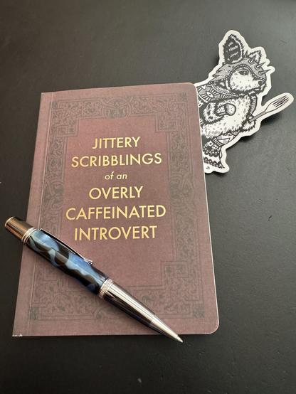 A journal lies on a black background.  It is patterned to resemble leather, but is paper.  The front is embossed (in gold print) with the legend "Jittery Scribblings of an Overly Caffeinated Introvert" and truer words were never written.

Tucked under the top the journal is my fav black-and-white sticker of "The Peckish Assassin" which I scored a few weeks ago at a local artist's spot in the #RAD in #Asheville.

Sitting on top of the journal is a chubby silver ball-point pen, with the barrel made out of a blue, black and white swirled acrylic.  My sister and her husband turn these acrylic blanks themselves and make the pens.