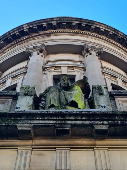 A statue of a seated female figures set in front of the columns of a Classical style portico.