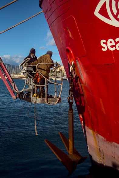 zwei perosnen in einer hebebühne vor dem bug eines schiffes, der anker ist ein stück herunter gelassen und baumelt kurz übers wasser