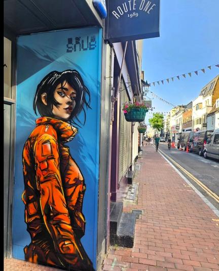 Streetartwall. A comic-style mural of a girl has been painted on a narrow exterior wall in the entrance area of a fashion store. A girl with wild, short black hair, fine facial features and a red aviator blouson is depicted on a blue background. She is looking over her shoulder at the viewer. The artist's name "Snub" is written at the top of the wall. The photo shows a view of the red sidewalk and the shopping street next to the mural.
Info: The mural is constantly being redesigned and has probably already been replaced by another figure by the artist.