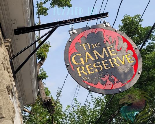 a round sign, with gold text and red images on a black background, depicting a dragon fighting a knight & wizard. Text reads "the game reserve"