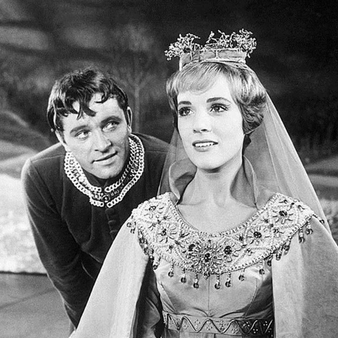 Black & white promotional shot of Camelot by Lerner & Loewe. Julie Andrews in a crown is seated, looking off in the distance. Richard Burton is crouching behind her, speaking almost in her ear.