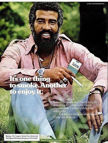 Atractive man smiles at the camera while sitting in a field enjoying a nice ciggy