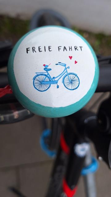 Fahrradklingel mit Aufschrift "FREIE FAHRT" über einem Damenrad, auf dessen Sattel ein Vogel Sitz und vor dem zwei rote Herzen fliegen