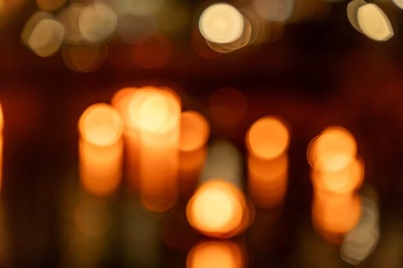 A color photograph, defocused  to the point that details have disappeared. Globes of orange and yellow light from flaming braziers on the water float above overlapping reflections caused by the water. Above, whiter lights from the surrounding city line the top of the frame, with a darker gap between the groups of lights.
