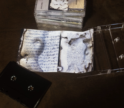 A black cassette in the foreground with the case opened on the table, insert opened to reveal bug eaten book pages. A stack of the same cassette visible in the background