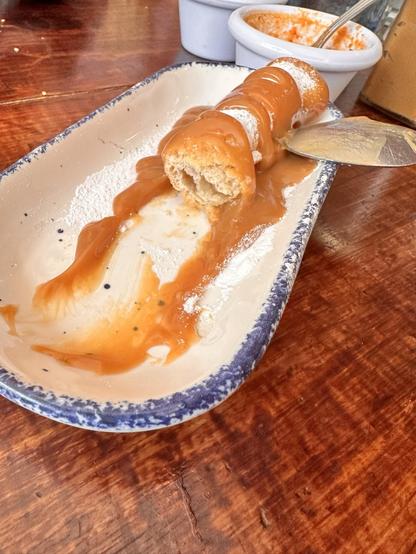 A partially eaten eclair with caramel sauce on a white plate with a blue rim, a knife, and a wooden table background.