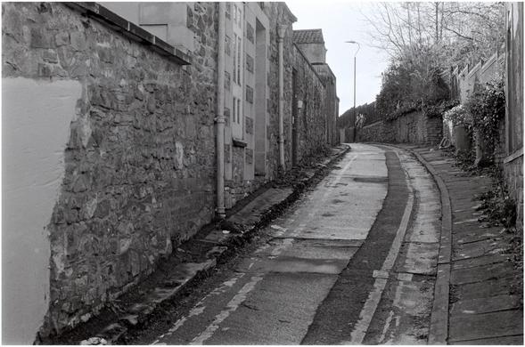 A black and white photograph taken in a wintry and wet Upper Church Lane off St Michael's Hill, Bristol.
Two bicycles are padlocked up on to railings in a narrow back lane.