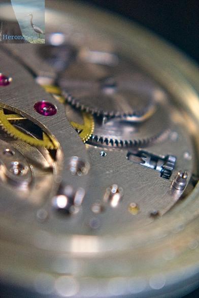A macro photograph of the internal gears and parts of a pocket watch, made of steel with brass gears and red ruby cylindrical bearings.
