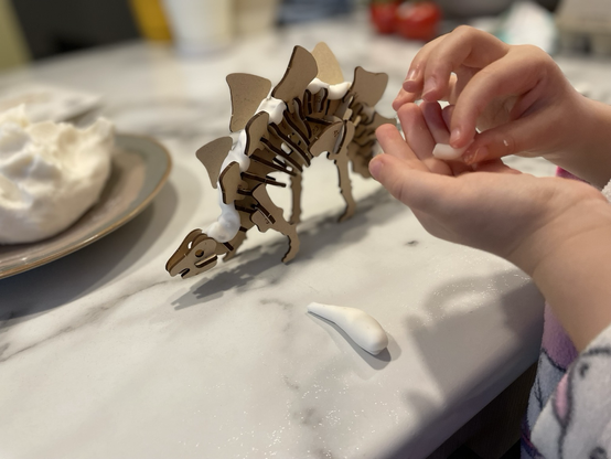 Small child hands shaping white modelling clay next to a wooden stegosaurus skeleton puzzle. The puzzle already has a strip of clay “muscle” along the spine.