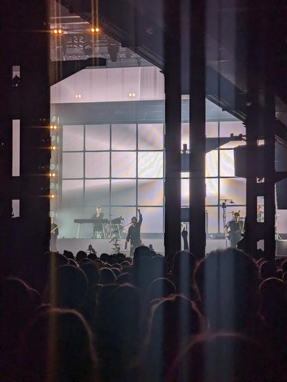 Foto durch Säulen hindurch von einer Bühne, deren Leinwand dahinter hell erleuchtet ist. Davor Silhouetten von Menschen an Instrumenten und einem Sänger in der Mitte