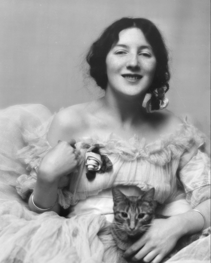 Portrait of early 20th-century art model and actress Audrey Muson. It is a formal studio portrait. She is dressed in a elaborate white gown and is hold a tabby cat on her lap. She is smiling and looking directly at the camera.