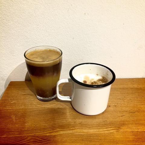 Ein Glas mit geschichtetem Espresso in Orangensaft und ein weißer Emaillebecher mit Milchschaum und Espresso auf einer hölzernen Oberfläche vor einer weißen Wand.
