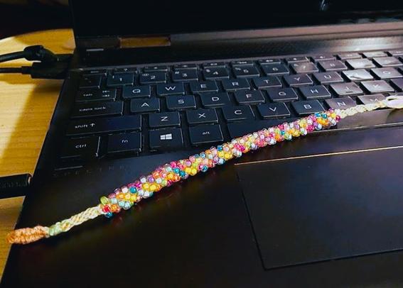 A black laptop keyboard with a kumihimo round braid with floss in pale orange and pale green, most of which is covered in small, candy-colored beads.