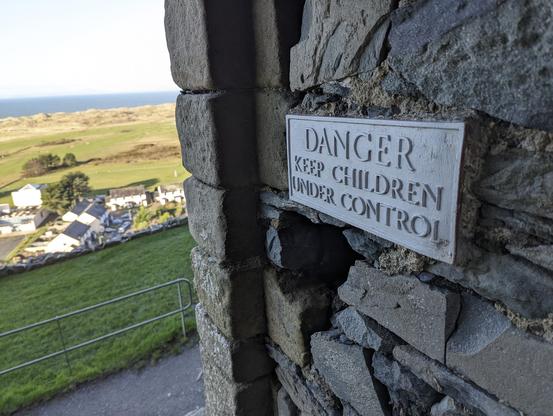 A sign affixed to a stone wall at Harlech Castle reads 'DANGER KEEP CHILDREN UNDER CONTROL', with a backdrop of golf course and the sea in the distance.