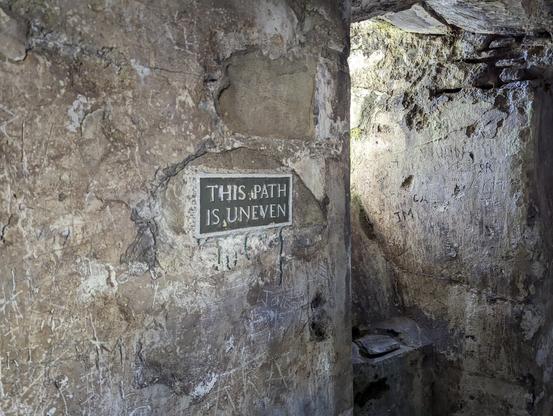 Inside Harlech Castle, a sign mounted on a rough stone wall states 'THIS PATH IS UNEVEN', surrounded by etchings and graffiti on the ancient walls.