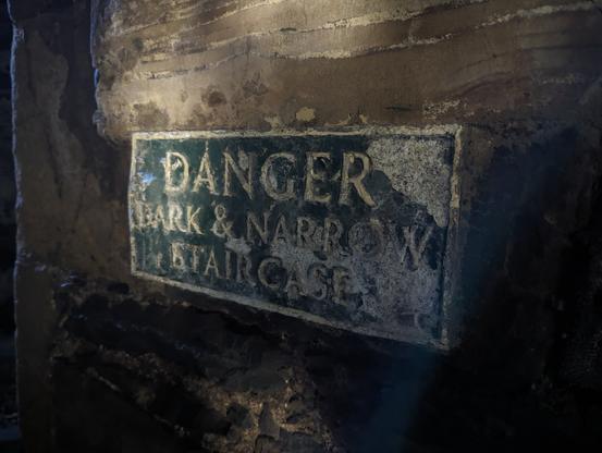 A weathered sign in Harlech Castle warns of 'DANGER DARK & NARROW STAIRCASE', set against a backdrop of dark, textured stone steps receding into shadow.