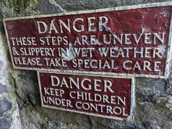 Two warning signs on a mossy stone wall at Harlech Castle: The upper sign is red with carved white letters saying 'DANGER THESE STEPS ARE UNEVEN & SLIPPERY IN WET WEATHER PLEASE TAKE SPECIAL CARE' and the lower sign mirrors the first with 'DANGER KEEP CHILDREN UNDER CONTROL'.