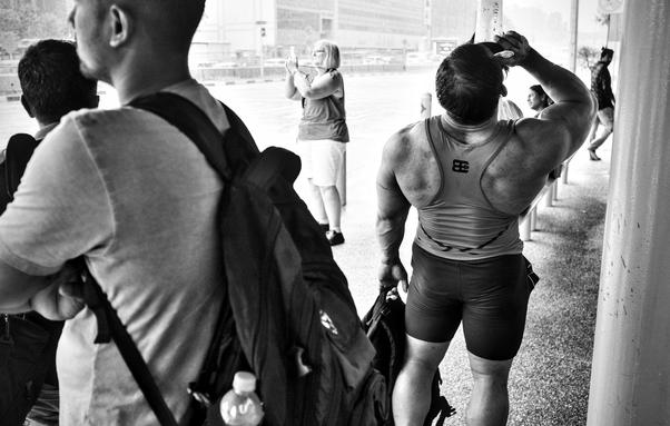 Passengers waiting at a bus stand, on the right is a body builder finishing his drink, middle a Caucasian woman taking a photo with her phone, on left passengers waiting