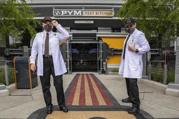 Two individuals in white lab coats and masks are standing in front of the "Pym Test Kitchen" entrance, with a trash can in the foreground. The left person is saluting, and the right person is giving a thumbs-up. The establishment appears to be a themed restaurant at a theme park.