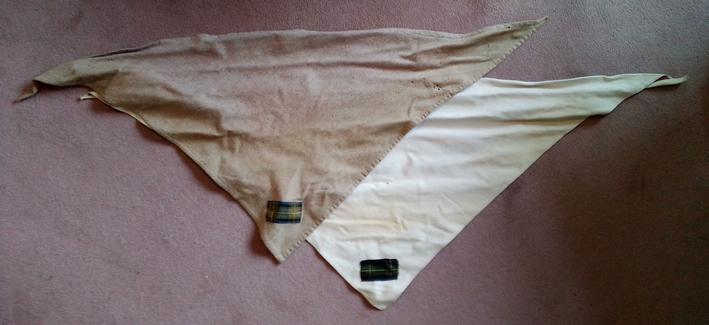 Two Scout scarves, triangular in shape. Different materials but very similar. Beige and stone in colour respectively. Each has a rectangular patch of predominantly green tartan in the corner.