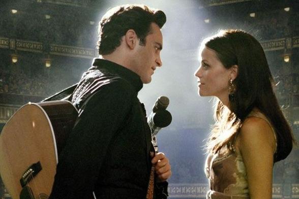 A man and woman stand on a stage in front of a big crowd. The man has a guitar on his back. There's a mic between the two people, but they're staring into each others' eyes.