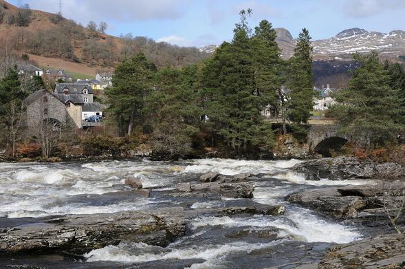 The impressive Falls of Dochart at Killin, with the River Dochart in full spate. The image shows the view down onto a broad expanse of rapids and rocks. Beyond then are trees and a stone bridge. There are buildings beyond the trees and in the background are mountains with some snow on them. The scene is in sunlight.