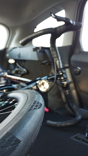 Ein schwarzes Rennrad liegt in einem Kofferraum. Im Vordergrund ist der Vorderreifen scharf zu sehen, das Rennrad und das Auto im Hintergrund sind unscharf.