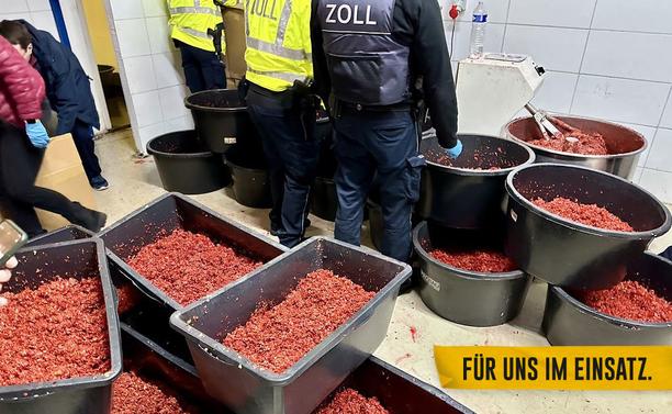 Einsatzkräfte des Zolls stehen in einer Produktionshalle für illegalen Wasserpfeifentabak. Um sie herum stehen mehrere Behälter mit rotem Wasserpfeifentabak.