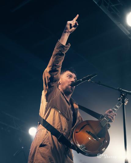 Robbie photographed from a low angle, wearing muted yellow overalls. His right hand is extended upwards, index finger pointing out, his left hand holds a mandolin by the neck, and he is visibly shouting something exciting into the microphone