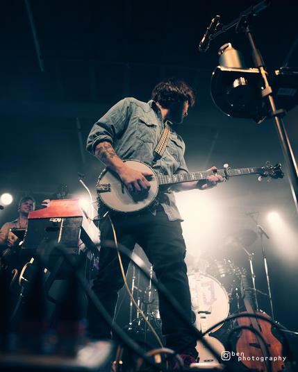 Andy photographed from a low angle, wearing a blue denim shirt and dark trousers. He is looking back over his left shoulder whilst in the middle of playing the banjo. In the background, where he's looking, are lots of white lights, a partially-visible drumkit, and off to the left you can see Antonio playing the upright bass