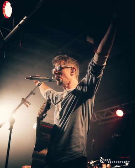 JD on a stage, wearing a grey long-sleeved t-shirt. He is backlit by misty white lights, both arms outstretched upwards with a guitar hanging freely from its strap, while he sings into the microphone