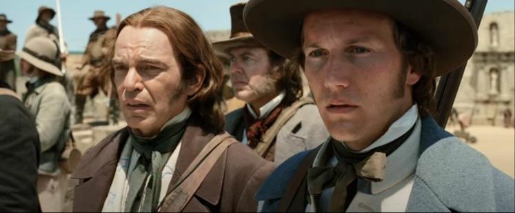 Two men in early American Victorian-era clothes and hair, with coats and high collars. They're standing amidst other men in various dress, some more rough and cowboy-style than others. Both men are looking out toward something offscreen and seem worried.