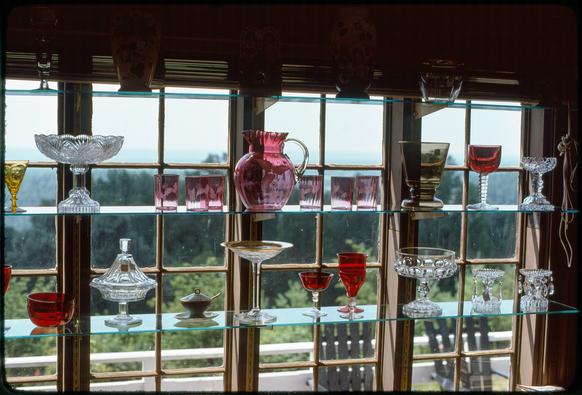 Various glassware—cup, vases, pitchers, goblets and bowls—sit on glass shelves in front of a multi-paned window. The glassware is of various colors and styles. Beyond the window is a small patio with a couple chairs. In the distance, the blurred background shows trees and a hazy ridge.