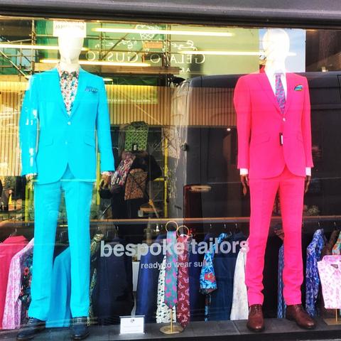 Two mannequins in a tailors window. The left one wearing a sky blue suit and the right one a bright pink suit.