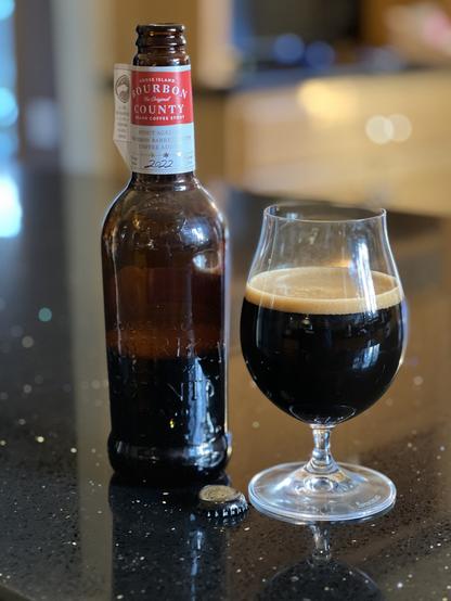 A bottle of Goose Island 2022 Bourbon County Coffee Stout with its iconic neck label with a red band and white lettering. A tulip glass is to the right half full of dark beer.