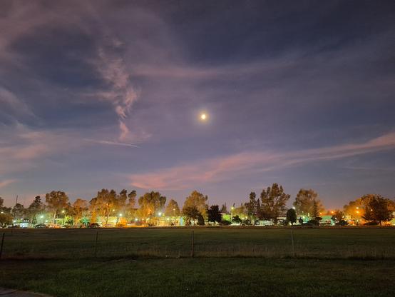 Night sky with clouds and the moon, the city lights on the horizon and some grass in the lower third of the photo