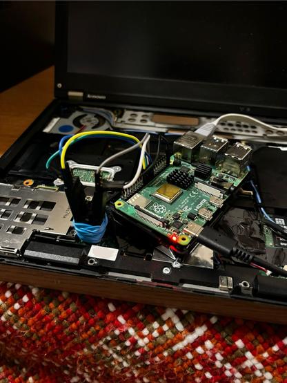 A photo of a laptop with the keyboard and wrist-rest removed, showing the motherboard. A Raspberry Pi is visible on top of the laptop with a 16-pin SOIC Clip connected to it. The clip itself is clipped to a chip on the motherboard and hold secure with blue rubber band.