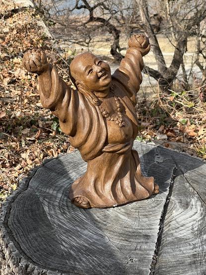 A laughing Buddha statue sips atop a stump, with a black walnut tree in the background. The Buddha is holding pineapples in both hands, reaching them toward the sky.