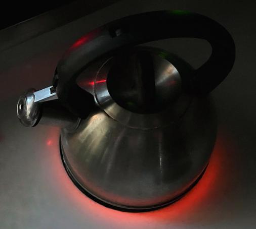 A metal tea kettle sitting on the stovetop burner in a darkened kitchen, the burner glowing red underneath the kettle as the water heats for tea.
