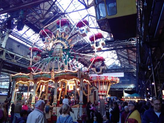 Innenansicht der belebten Jahrhunderthalle Bochum, ein altes Industriegebäude. vorne ein altes Riesenrad, dahinter ist noch ein Kettenkarussell sichtbar.